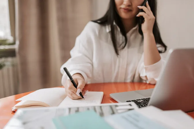 Someone on a phone call, writing notes in a notebook at a desk
