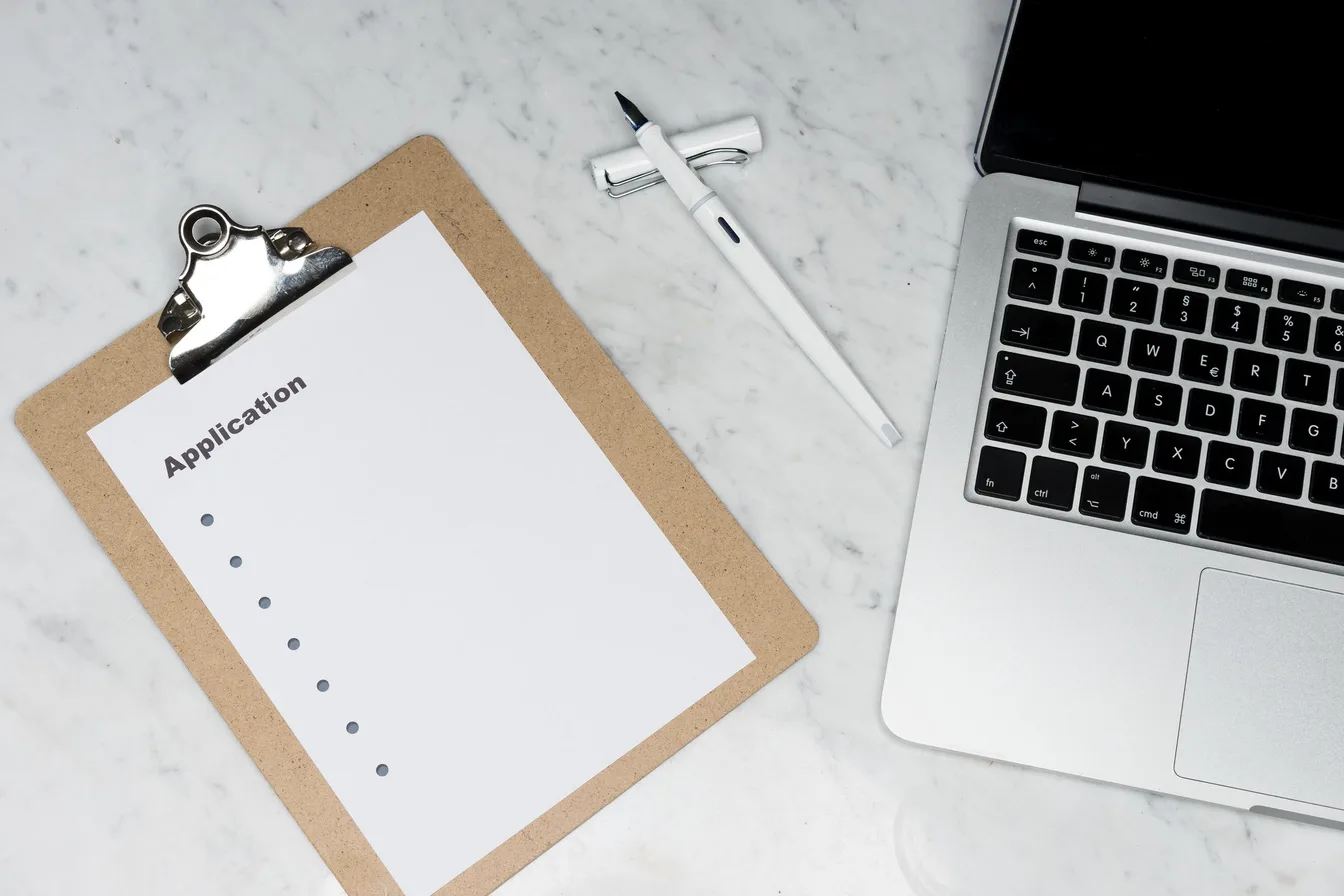 A clipboard with a blank application form beside a pen and a laptop